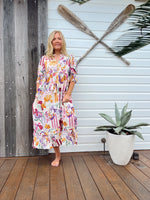 Woman in a colorful dress standing on a wooden deck with a plant and oars in the background.