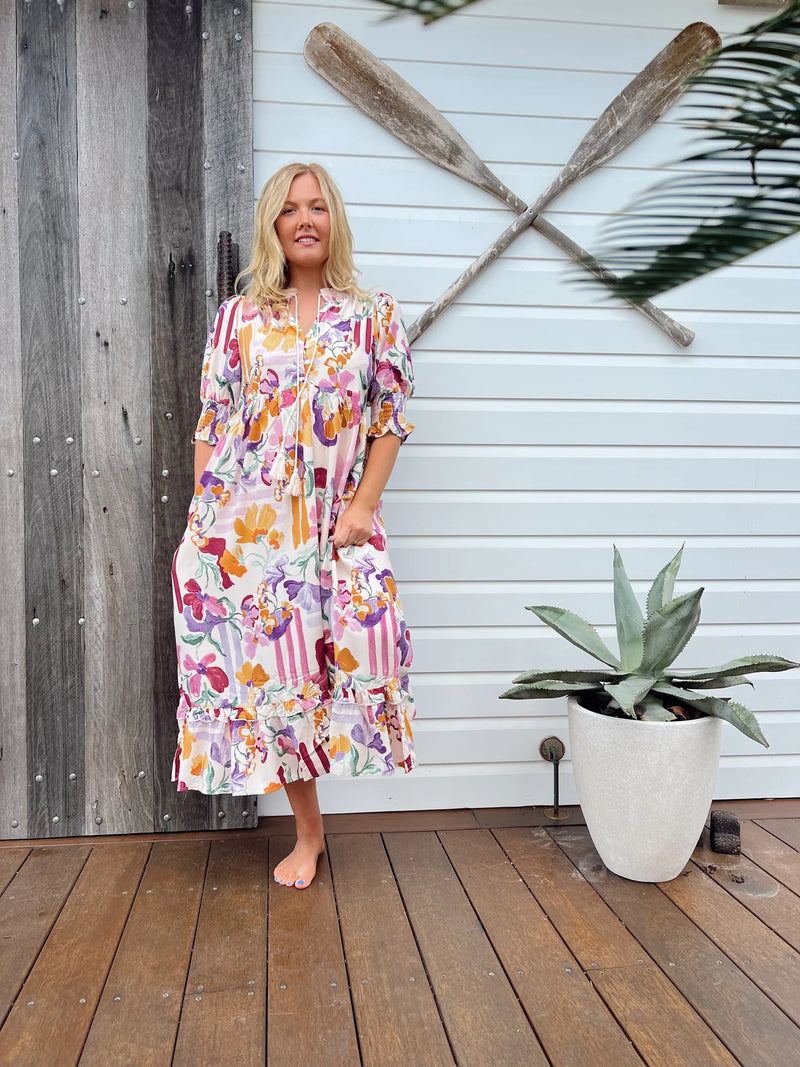 Woman in a colorful dress standing on a wooden deck with a plant and oars in the background.