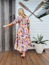 Woman in a colorful floral dress standing on a wooden deck with oars and a plant in the background.