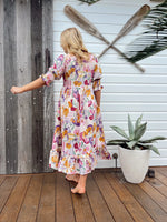 Woman in a colorful floral dress standing on a wooden deck with oars and a plant in the background.