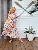 Woman in a floral dress standing on a wooden deck with oars and a plant in the background.