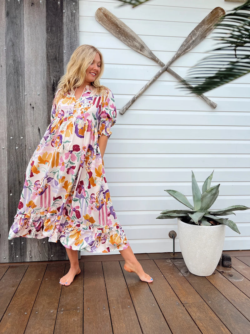 Woman in a floral dress standing on a wooden deck with oars and a plant in the background.