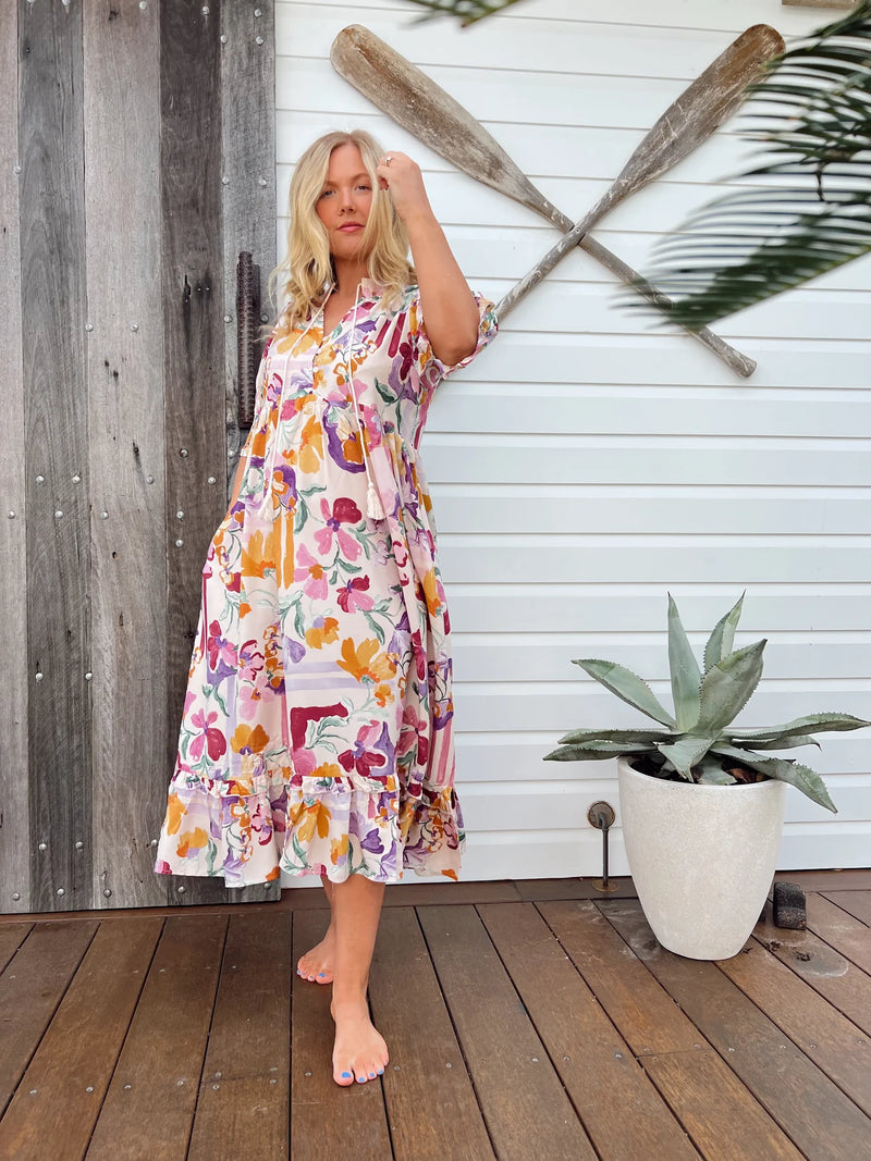 Woman in a floral dress standing on a wooden deck with oars and a plant in the background.