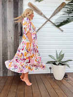 Woman in a colorful floral dress standing on a wooden deck with oars and a plant in the background.