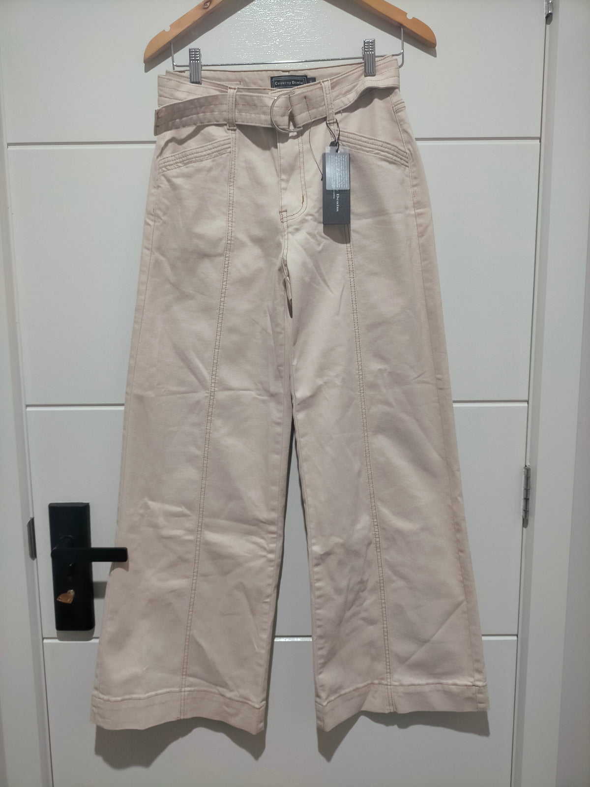 Beige pants hanging on a wooden hanger against a white tiled wall.