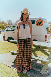 Jaase Stevie Austin Flannel Pants chocolate by Jaase – model standing in front of beach park bench table looking at camera with cowboy hat white cami and thongs outdoor lifestyle shot