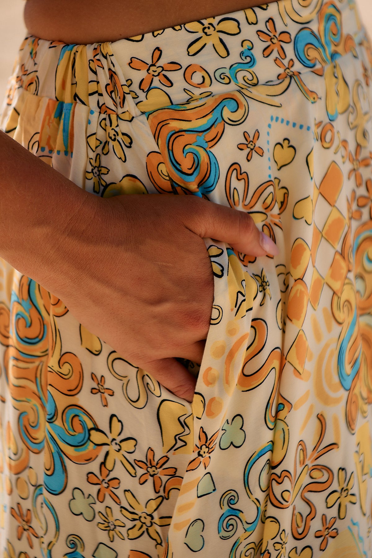 Close-up of a person wearing the Sonnie print which is a buttery yellow colorful patterned maxi skirt with a hand showing it has pockets.